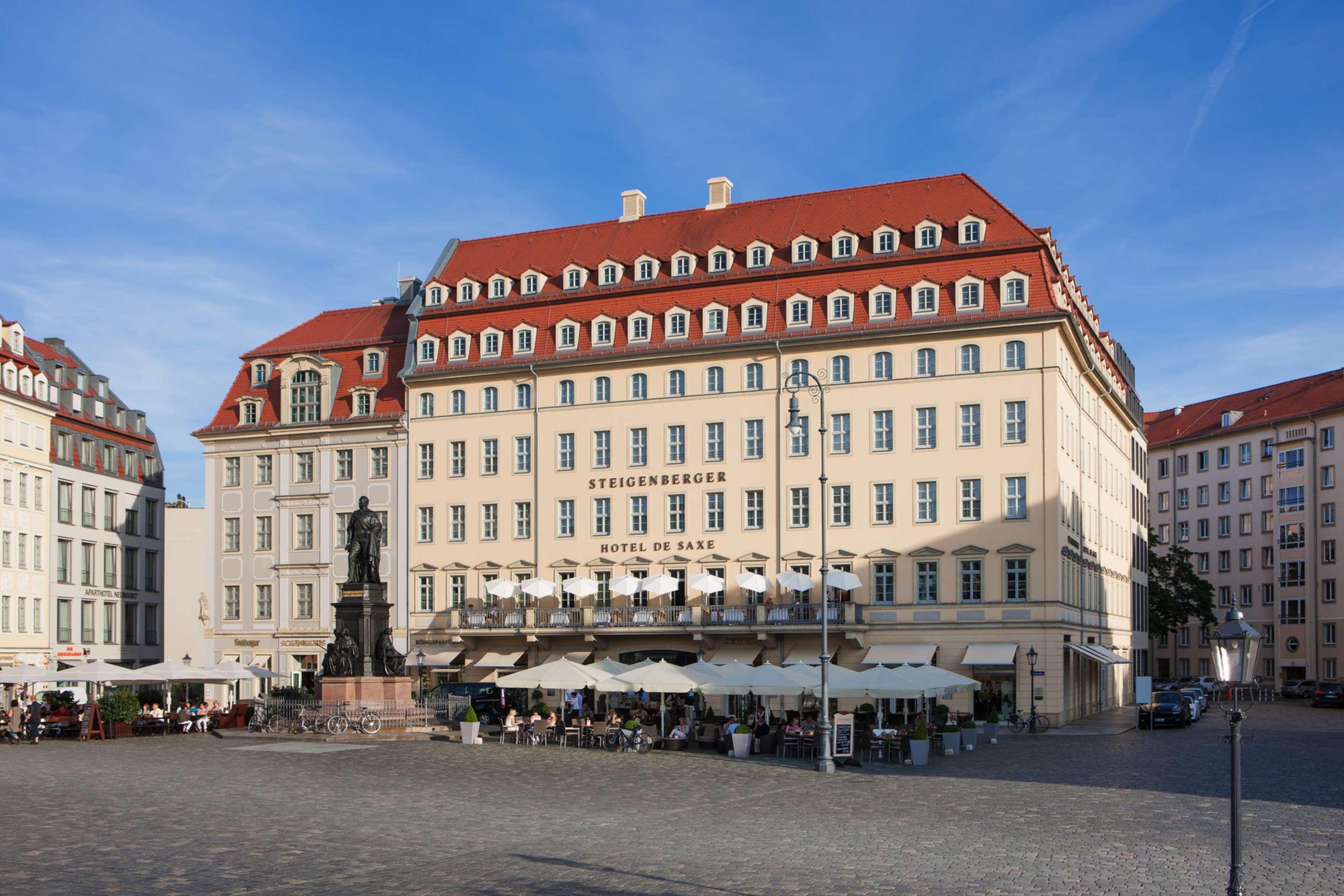 Steigenberger Hotel De Saxe Dresden Exterior photo The photo shows a large, elegant building with a light-colored facade and a distinct red roof. The structure features multiple windows and a decorative style typical of historic architecture. In front of the building, there is a plaza with outdoor se